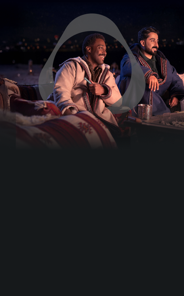 رجلان يجلسان في الهواء الطلق ليلاً، يرتديان عباءات تقليدية دافئة، يبتسمان ويستمتعان بجلسة مريحة مع وسائد مزخرفة وطاولة تحتوي على شاي ووجبات خفيفة.