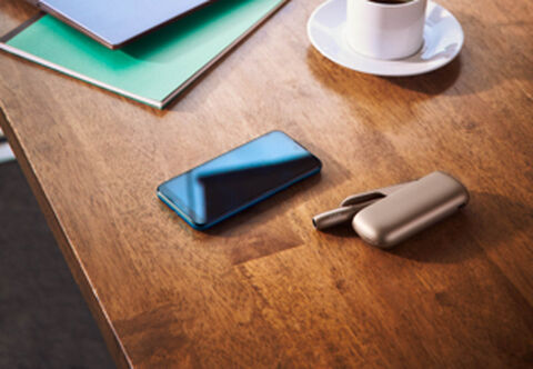 A gold IQOS, a mobile and a cup of coffee on a wooden table.