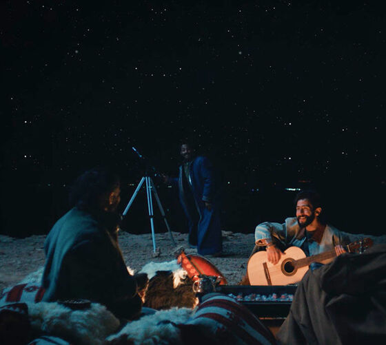 Group of friends sitting outdoors at night under a starry sky, one playing guitar by the fire, another near a telescope, enjoying a cozy gathering.
