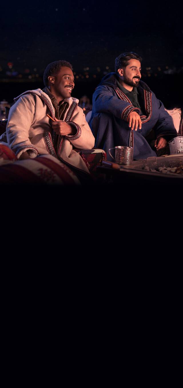 Two men sitting outdoors at night, dressed in traditional warm cloaks, smiling and enjoying a cozy gathering with patterned cushions and a table with tea and snacks.