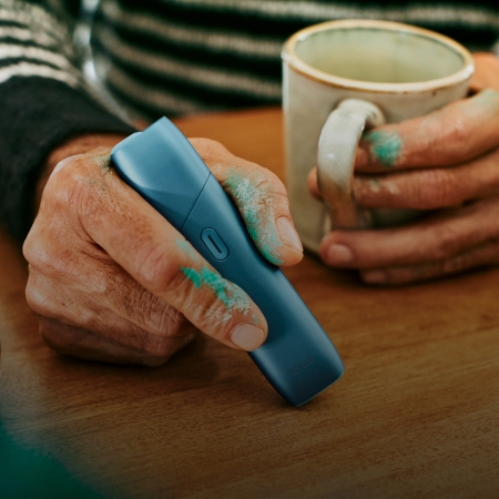Hands holding a coffee cup and a turquoise IQOS Originals One heated tobacco device.