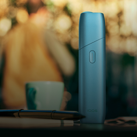 Turquoise IQOS Originals One device on a coffee table next to coffee cup.