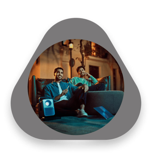 Two young men sitting on sofas outdoors at night, smiling and relaxing with an IQOS device and a portable projector nearby.