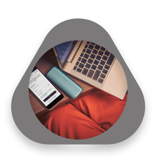 Smartphone with IQOS app open next to a turquoise IQOS device and a laptop on a wooden table, with a person in red pants sitting nearby.
